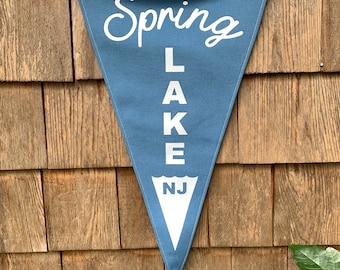 May include: A blue triangular pennant with white text that reads "Spring Lake NJ". The pennant has a tan fabric border and is hanging on a wooden wall.