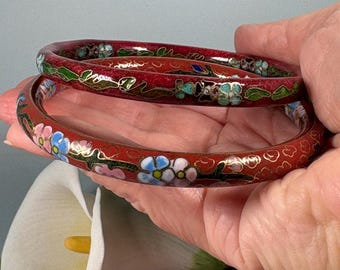 May include: Two circular red bangles featuring intricate cloisonné enamel work. The bracelets are decorated with colorful floral patterns in shades of blue, pink, and green, outlined with thin gold-toned metal wire detailing.