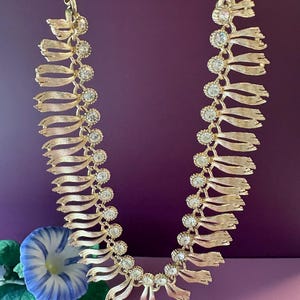 May include: Gold-toned necklace with a ribbon design, adorned with clear, round gemstones. The necklace features a series of ribbon-like elements connected by the gemstones. The background is a solid purple color, with a blue and white flower in the lower left corner.