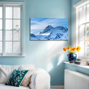 Könnte beinhalten: Gerahmter Landschaftsdruck mit schneebedeckten Bergen unter blauem Himmel mit Wölkchen. Der Druck hängt an einer hellblauen Wand über einem weißen Sofa. Eine blaue Vase mit orangefarbenen Blumen steht auf einer Fensterbank.
