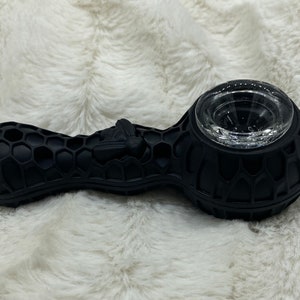 May include: Black silicone pipe with a honeycomb pattern and a clear glass bowl. A bee is sculpted on the side. The pipe is designed for smoking and rests on a white, fluffy surface.
