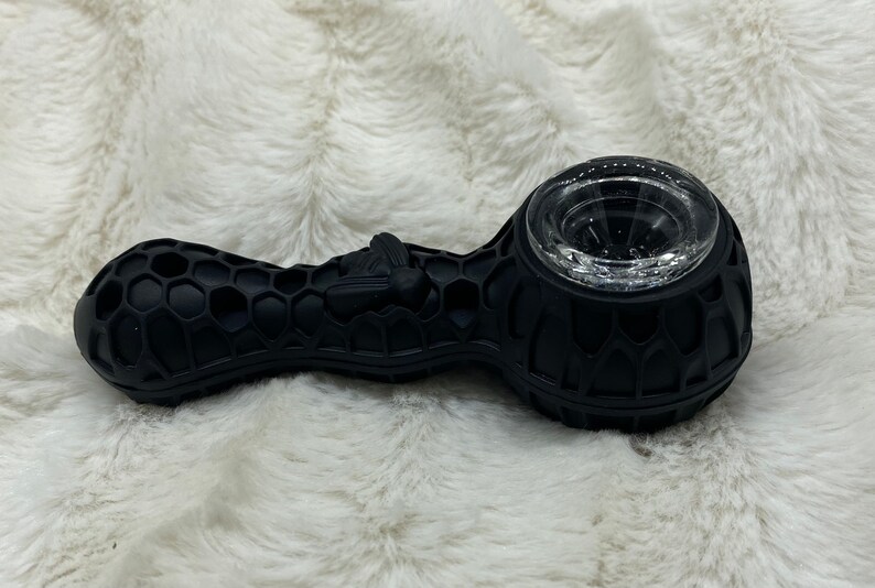 May include: Black silicone pipe with a honeycomb pattern and a clear glass bowl. A bee is sculpted on the side. The pipe is designed for smoking and rests on a white, fluffy surface.