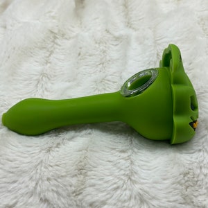 May include: A green silicone smoking pipe with a skull-shaped bowl. The pipe has a clear glass bowl and a long, curved stem.
