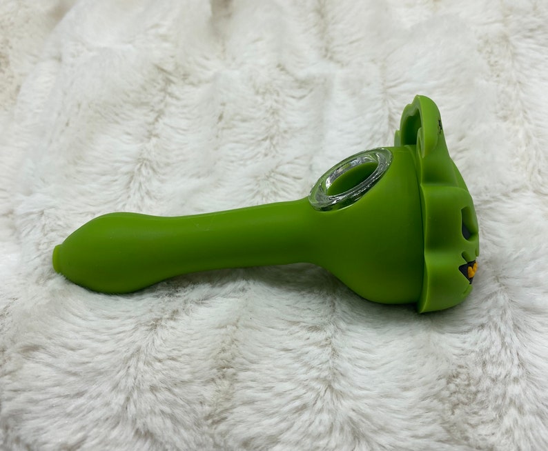 May include: A green silicone smoking pipe with a skull-shaped bowl. The pipe has a clear glass bowl and a long, curved stem.
