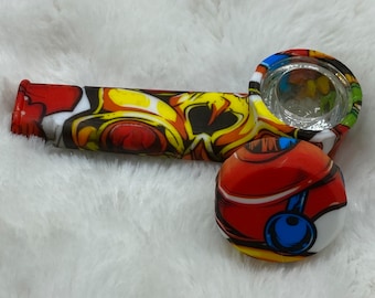 May include: A colourful silicone pipe with a glass bowl. The pipe features a red, yellow, and white design. The bowl is clear, and the mouthpiece is red. The pipe is designed for smoking.