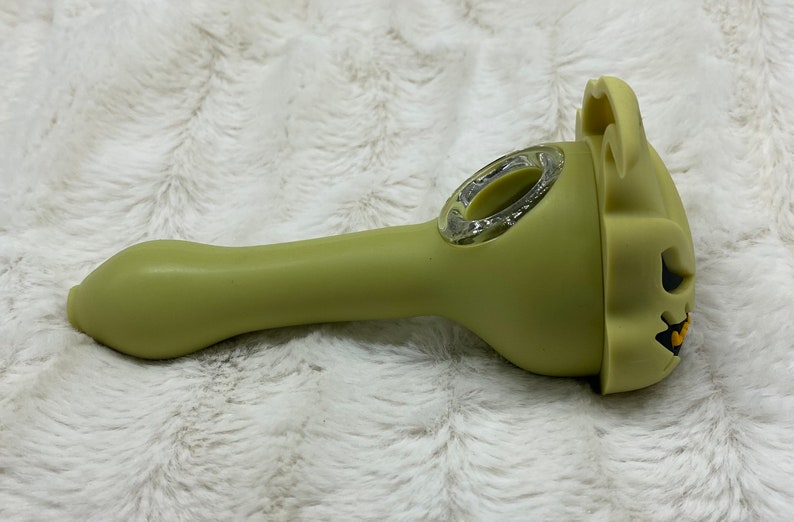 May include: A green silicone smoking pipe shaped like a jack-o'-lantern with a clear glass bowl.