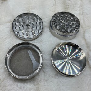 May include: A four-piece metal grinder with a silver finish. The grinder has a top piece with a series of holes, a middle piece with teeth, a bottom piece with a screen, and a separate scraper tool.