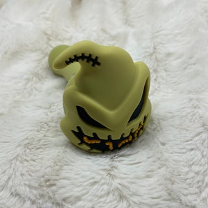 May include: Green silicone pipe shaped like the character Oogie Boogie from the movie The Nightmare Before Christmas. The pipe has a stitched detail on the top and a black and yellow face with a stitched mouth.