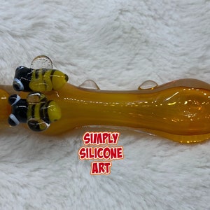 May include: A honey-colored glass pipe with three bee-shaped glass accents. The pipe is decorated with black and yellow stripes and white accents. The text "SIMPLY SILICONE ART" is visible below the pipe.