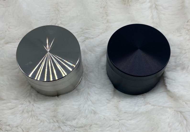 May include: Two metal grinders, one silver with a starburst design and one black. Both grinders have a textured surface.