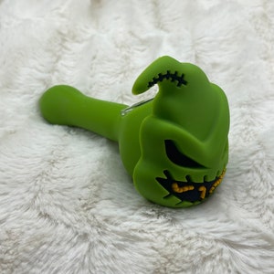 May include: Green silicone smoking pipe shaped like the character Jack Skellington from the movie The Nightmare Before Christmas. The pipe has a black stitched mouth and yellow eyes.