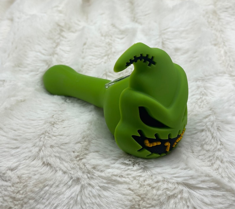 May include: Green silicone smoking pipe shaped like the character Jack Skellington from the movie The Nightmare Before Christmas. The pipe has a black stitched mouth and yellow eyes.