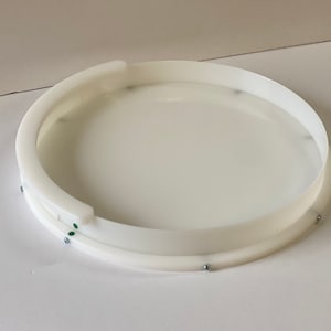 May include: White plastic circular tray with a raised edge and a smaller inner ring. The tray has several small silver screws around the edge.