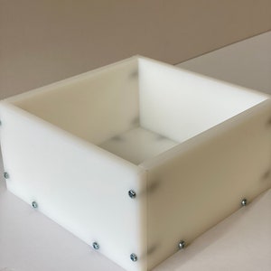 May include: A white plastic box with a square shape and a smooth surface. The box is assembled with screws visible on the sides.