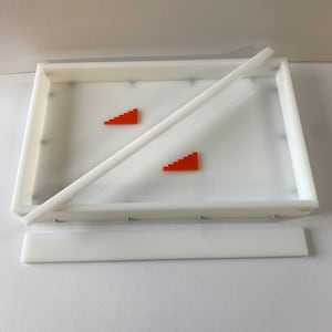 May include: A white rectangular tray with a clear interior, featuring two orange stair-step shapes. A white bar rests diagonally across the tray, with a detached white rectangular piece below. The tray is designed for a specific purpose.