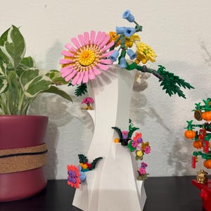 May include: A white, geometric vase with colourful, artificial flowers. The arrangement includes a large pink daisy, yellow and blue flowers, and green foliage. The vase is decorated with smaller flowers.