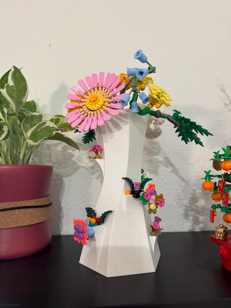 May include: A white, geometric vase with colourful, artificial flowers. The arrangement includes a large pink daisy, yellow and blue flowers, and green foliage. The vase is decorated with smaller flowers.