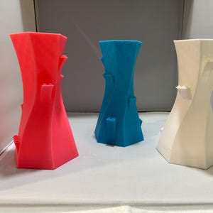 May include: Three decorative vases in red, blue, and white. Each vase has a unique twisted design with small, rectangular protrusions. The vases are displayed against a neutral background, showcasing their vibrant colours and modern aesthetic.