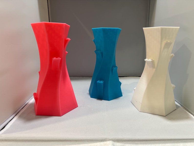 May include: Three decorative vases in red, blue, and white. Each vase has a unique twisted design with small, rectangular protrusions. The vases are displayed against a neutral background, showcasing their vibrant colours and modern aesthetic.