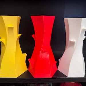 May include: Three abstract vases in yellow, red, and white. Each vase has a unique, twisted design with architectural elements. The vases are displayed on a black surface against a dark background, showcasing their vibrant colours and modern aesthetic.
