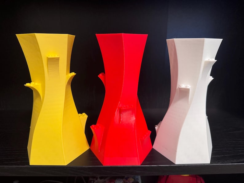 May include: Three abstract vases in yellow, red, and white. Each vase has a unique, twisted design with architectural elements. The vases are displayed on a black surface against a dark background, showcasing their vibrant colours and modern aesthetic.