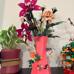 May include: A bright pink vase displays a bouquet of colourful flowers, including large magenta orchids and peach-coloured blooms. The vase is decorated with small, decorative flower arrangements. A potted plant and a miniature tree are visible in the background.