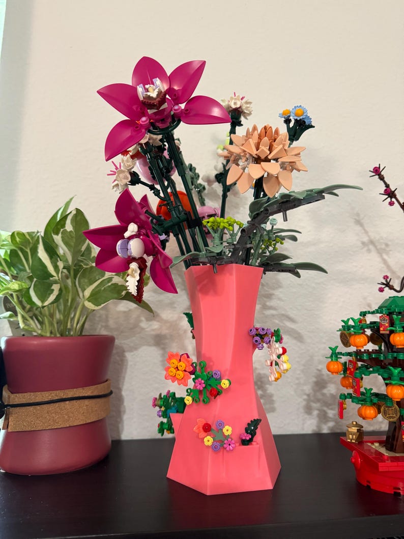 May include: A bright pink vase displays a bouquet of colourful flowers, including large magenta orchids and peach-coloured blooms. The vase is decorated with small, decorative flower arrangements. A potted plant and a miniature tree are visible in the background.