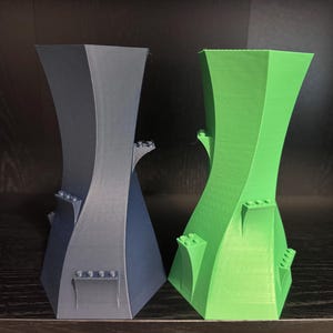 May include: Two abstract, 3D-printed vases. One is dark grey, the other is bright green. Both have a unique, curved design with small, rectangular details. The vases are displayed on a dark surface.