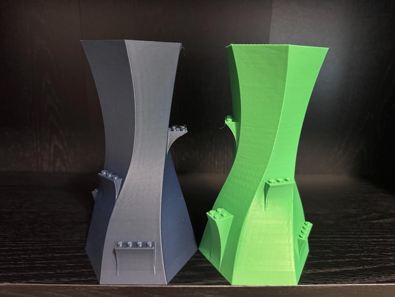 May include: Two abstract, 3D-printed vases. One is dark grey, the other is bright green. Both have a unique, curved design with small, rectangular details. The vases are displayed on a dark surface.