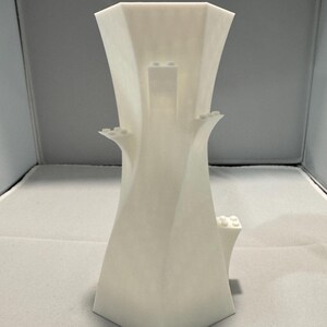 May include: A white, geometric vase with an unusual, architectural design. The vase has a flared top and small, rectangular protrusions. The shape is reminiscent of a modern sculpture. The vase is approximately 20 cm tall.