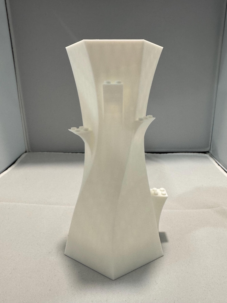 May include: A white, geometric vase with an unusual, architectural design. The vase has a flared top and small, rectangular protrusions. The shape is reminiscent of a modern sculpture. The vase is approximately 20 cm tall.