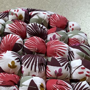 Könnte beinhalten: Ein runder, gesteppter Kissen mit einem Blumenmuster in Rot, Weiß und Burgunder. Die Oberseite besteht aus einzelnen, aufgeplusterten Quadraten. Die Seiten zeigen ein sich wiederholendes Muster aus Blättern und Blumen.