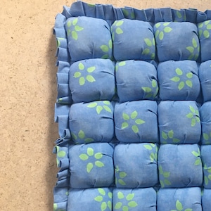 May include: A blue and green patterned floor cushion with a ruffled edge. The cushion is made of fabric and is divided into squares.