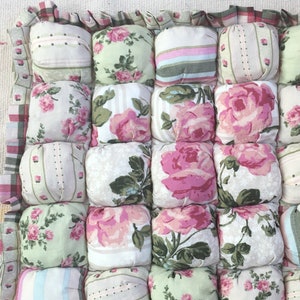 May include: A square cushion with a patchwork design featuring various floral patterns in shades of pink, green, and white. The cushion is made of fabric and has a ruffled border.