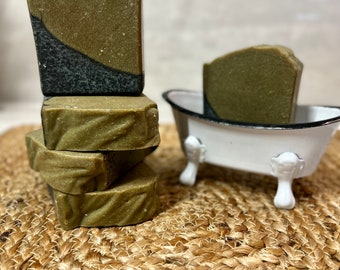 May include: A stack of olive green soap bars with a dark gray accent. One bar rests in a miniature white bathtub with claw feet. The soaps are on a woven, circular mat.