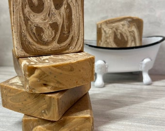 May include: Four bars of handmade soap stacked on top of each other. The soap is brown and white with swirls and flecks of brown. The soap is sitting on a light wood surface.