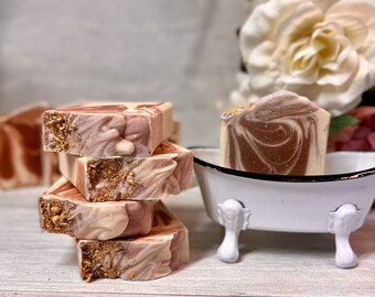 May include: Four bars of handmade soap stacked on top of each other. The soap is a swirl of brown, white, and pink with gold flecks. The soap is sitting on a light wood surface. A small white bathtub with black trim is in the background.