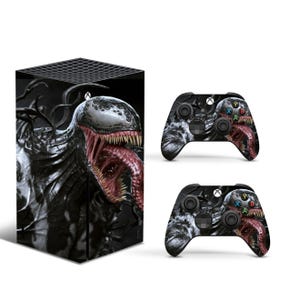 May include: A black and white gaming console and two matching wireless controllers featuring a graphic print of the character Venom with a wide mouth, sharp teeth, and a long red tongue. The console has a square shape with a black grid top.