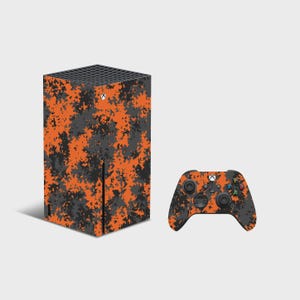 May include: A rectangular gaming console and a wireless controller featuring a vibrant orange and dark gray digital camouflage pattern. The console has a black top vent, a circular power button, and a vertical slot. The controller has black buttons.
