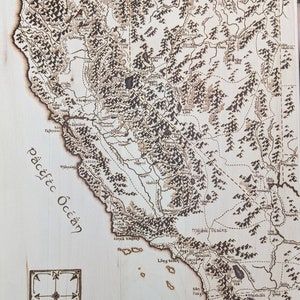 Puede incluir: Un mapa de California quemado en madera con el Océano Pacífico, montañas y lagos etiquetados. El mapa está sobre un fondo de madera clara e incluye una rosa de los vientos en la esquina inferior izquierda.