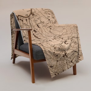 May include: A tan throw blanket draped over a gray armchair. The blanket features a detailed map design in shades of brown, with intricate lines and text. The chair has wooden arms and legs, set against a neutral background.