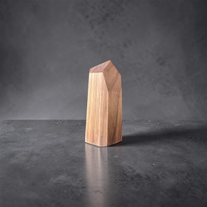 May include: A wooden obelisk sculpture with a geometric design. The light brown wood has visible grain and a faceted top. The sculpture stands on a dark grey surface against a matching background.