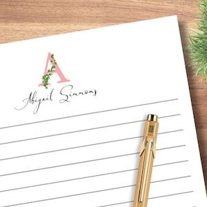 May include: A pink letter A with green vines and white flowers, with the name Abigail Simmons written in cursive below.