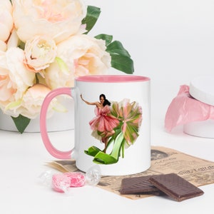 Puede incluir: Una taza de cerámica blanca con un borde y un asa de color rosa. La taza presenta una ilustración colorida de una mujer con un vestido rosa y zapatos verdes. El fondo es blanco.
