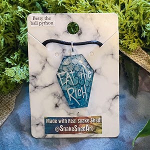 May include: A blue and white pendant necklace with the words "Eat the Rich" in white lettering. The pendant is coffin-shaped and hangs from a black cord. The necklace is displayed on a marble-patterned card with text that reads "Betty the ball python" and "Made with Real Snake Shed! @SnakeShedArt".