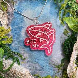 May include: A pink and white shark charm necklace with the text "BITE ME" on the charm.