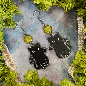 May include: A pair of black cat-shaped earrings with bat wings and white accents. The earrings have silver-colored hooks and are adorned with round, yellow-green cabochons. The earrings are set against a backdrop of green moss and a blue-toned background.