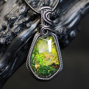 Könnte beinhalten: Ein handgefertigter Anhänger mit einem von der Natur inspirierten Design. Der tropfenförmige Anhänger hat ein lebendiges grünes und gelbes Harz mit eingebetteten Naturelementen, das in einem silberfarbenen Drahtrahmen eingefasst ist. Der Anhänger hängt an einer schwarzen Kordel.