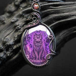 Könnte beinhalten: Ein ovaler Anhänger mit einer violetten Illustration einer Katze mit einer Mondsichel auf der Stirn, umgeben von Kerzen und Symbolen. Das Bild ist in einen dunklen, oxidierten, drahtumwickelten Metallrahmen mit einer schillernden Perle gefasst.