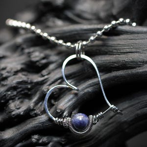 May include: A silver heart-shaped pendant necklace with a blue gemstone bead. The necklace is on a dark gray background.
